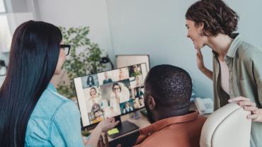 Drei Mitarbeitende stehen bzw. sitzen um einen Bildschirm, auf dem eine Videokonferenz zu sehen ist.