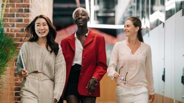 Drei junge Frauen in Businesskleidung laufen lachend einen Flur hinunter.