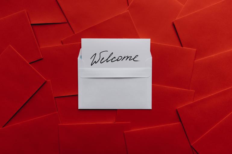 Viele rote Briefumschläge, in der Mitte ein weißer aus dem eine weiße Karte mit der Aufschrift "Welcome" herausguckt.
