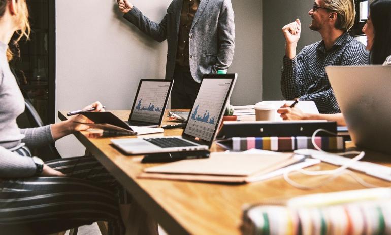 Foto: Ronald Candonga / Pixabay Büroszene: Schreibtisch mit Laptops und Arbeitsunterlagen, im Hintergrund schreibt jemand an ein Whiteboard.