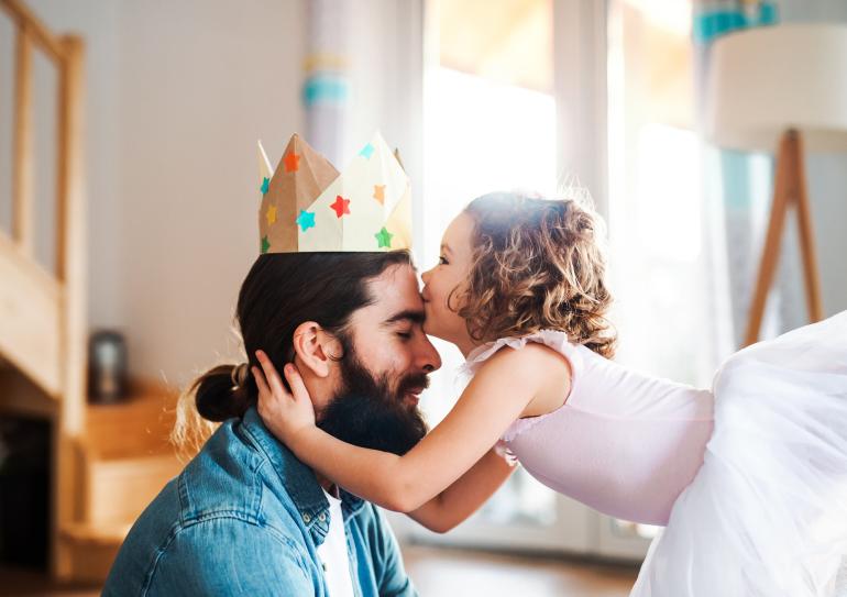 Ein Vater mit einer gebastelten Krone auf dem Kopf erhält von seiner kleinen Tochter im Tütü einen Kuss auf die Stirn.
