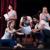 Eine Gruppe junger Schauspielerinnen und Schauspieler sitzt und steht auf der Bühne und geht die Texte durch.