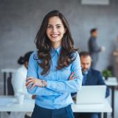 Foto: Dragana Gordic - Adobe Stock Eine junge Frau in Businesskleidung steht vor einem Bürohintergrund.