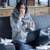 Foto: Monstar Studio - Adobe Stock Eine schwarzhaarige Frau sitzt vor einem Laptop auf dem Sofa und putzt sich die Nase, in der Hand hat sie eine Teetasse.
