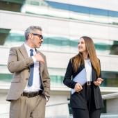 Foto: Vadim Pastuh - Adobe Stock Ein mittelalter Mann und eine junge Frau in Businesskleidung laufen nebeneinander, im Hintergrund sieht man ein Bürogebäude.